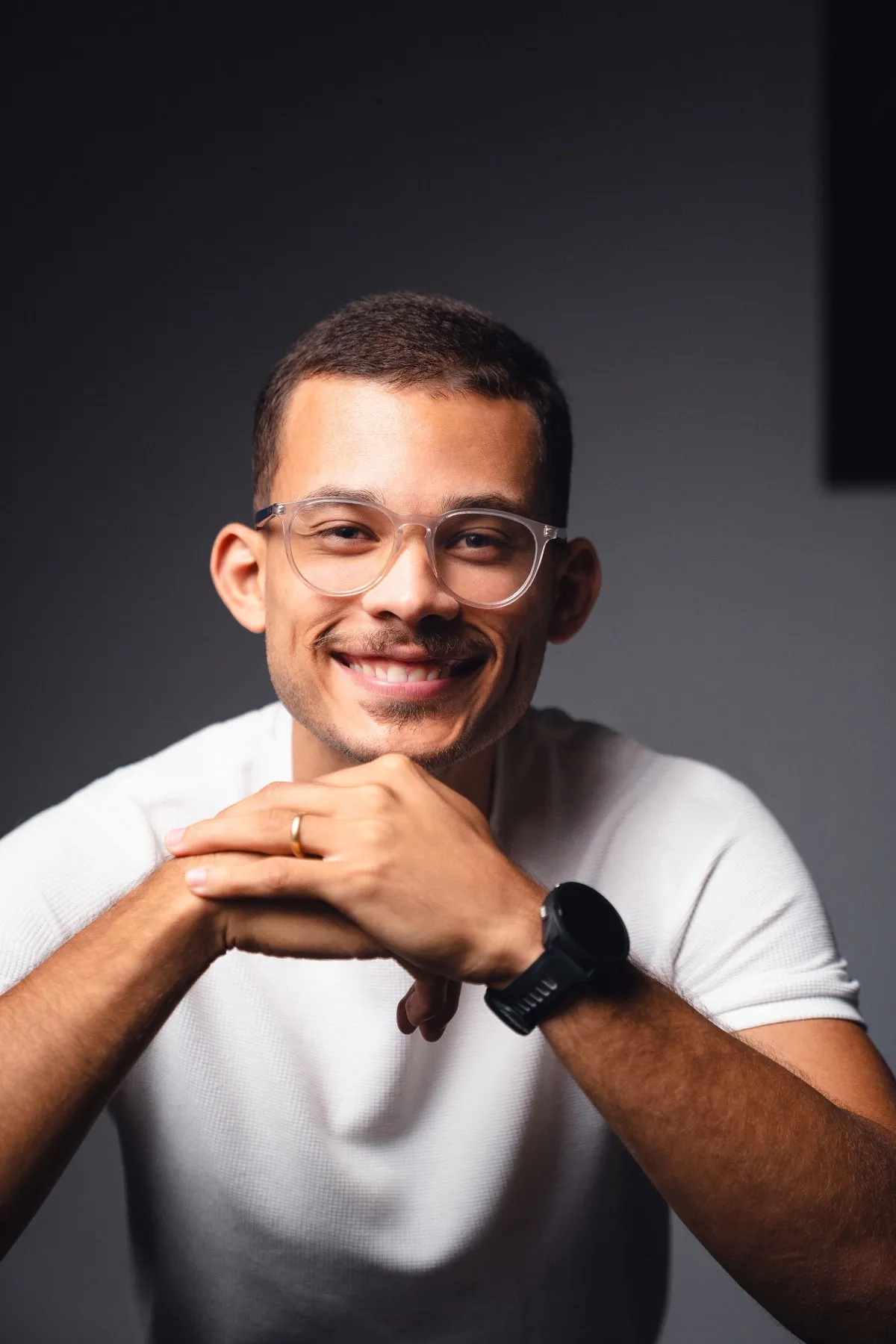 Marlon Torres sorrindo, de óculos e camiseta branca, com as mãos entrelaçadas